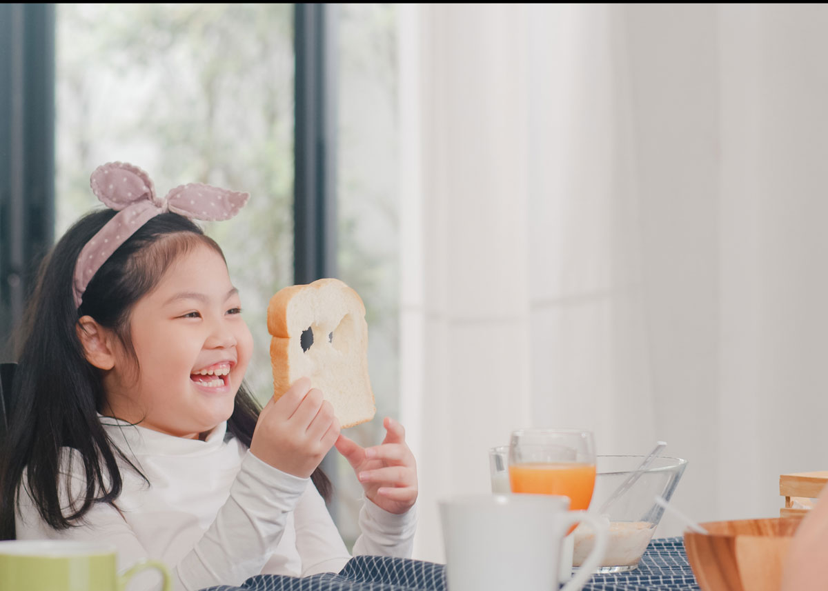Little girl sat at the kitchen table holding up a piece of bread which she's poked holes in, laughing at it.