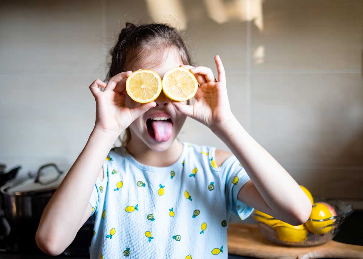 Girl wearing a top with lemon print on it, holding up two halves of a lemon as bug eyes and sticking her tongue out.