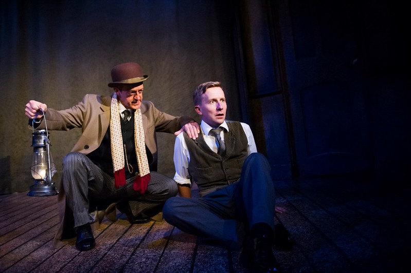 The two actors in the show The Women in Black on stage, one sitting down looking terrified and the other leaning his hand on his shoulder holding a lantern.