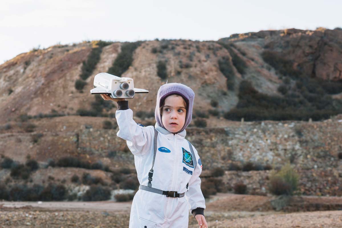 Little child dressed as an astronaut standing outside on rocky hills making their toy rocket fly.