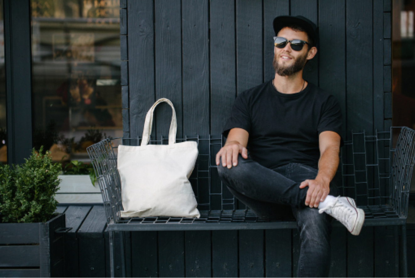 A guy sitting next to his tote bag on a bench.