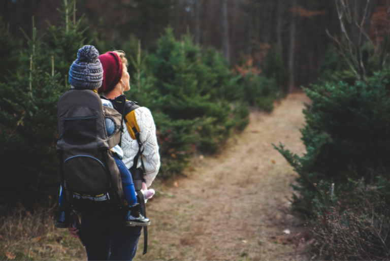 A woman carries a baby on her back as she walks through a clearing in a forest
