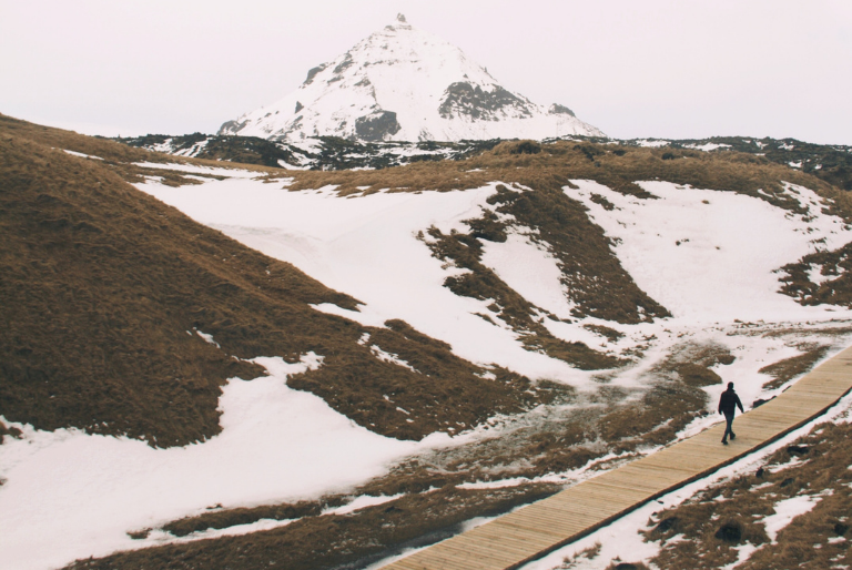 A person walks along a boardwalk in a snowy landscapes and mountain in the distance