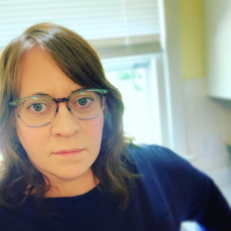 Portrait of Rachael Dietkus. She's wearing a dark longsleeve and glasses. Behind her is a window.