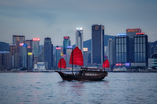 a boat with red ribbed sails on a river with Hongkong cityscape in the background