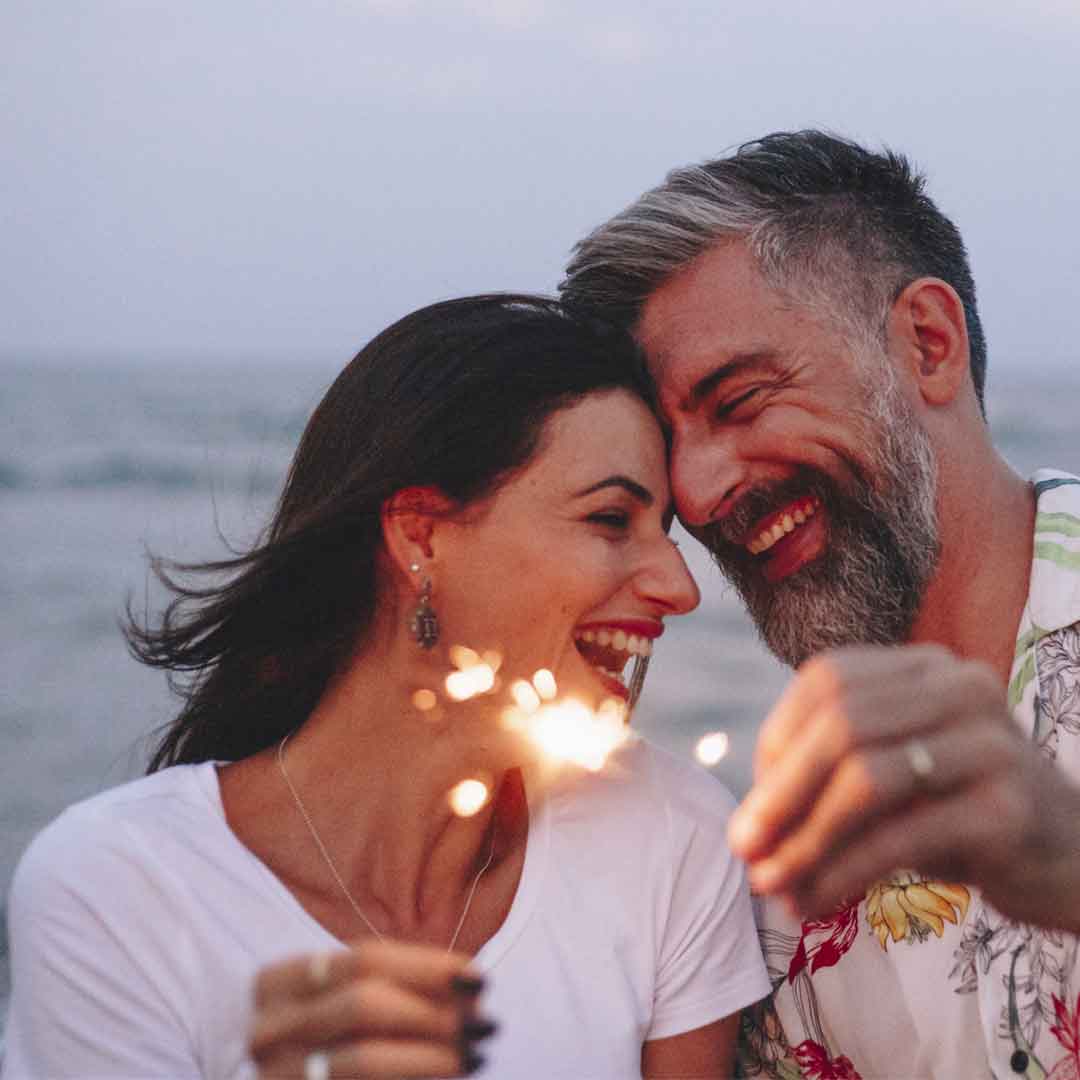a mature couple lean their smiling faces together while holding sparklers on the beach at twilight