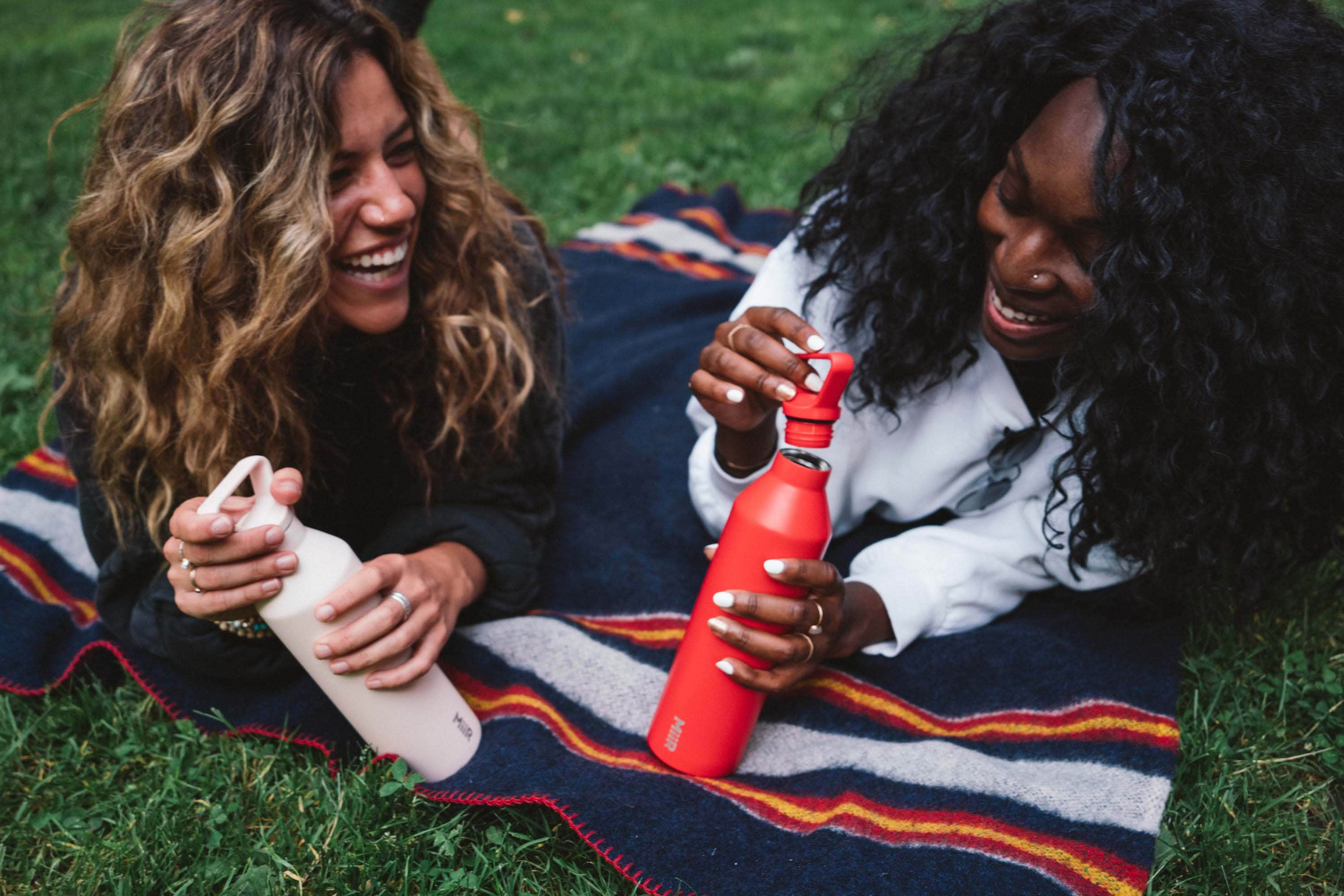 Two women lounge on a picnic blanket. Both hold MiiR bottles.
