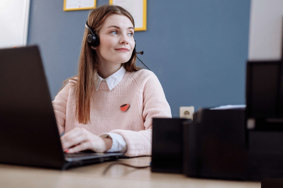 A girl with a headset, sitting at a desk with her laptop open, hopefully looking to the side