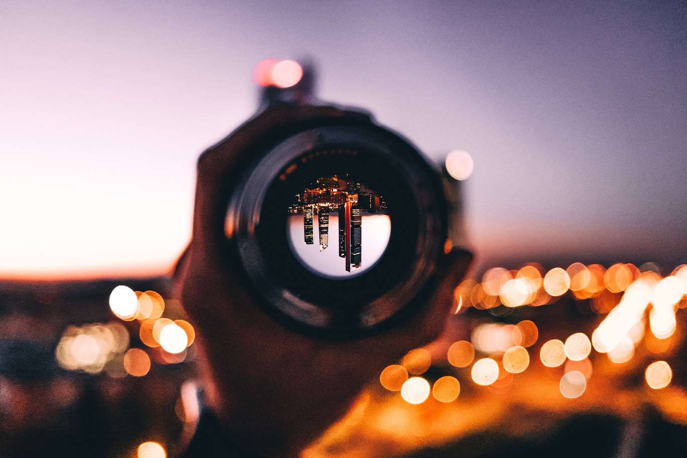 A blurred, nighttime cityscape in the background and a person holding up a telescope lens that shows an upside-down view of skyscrapers.
