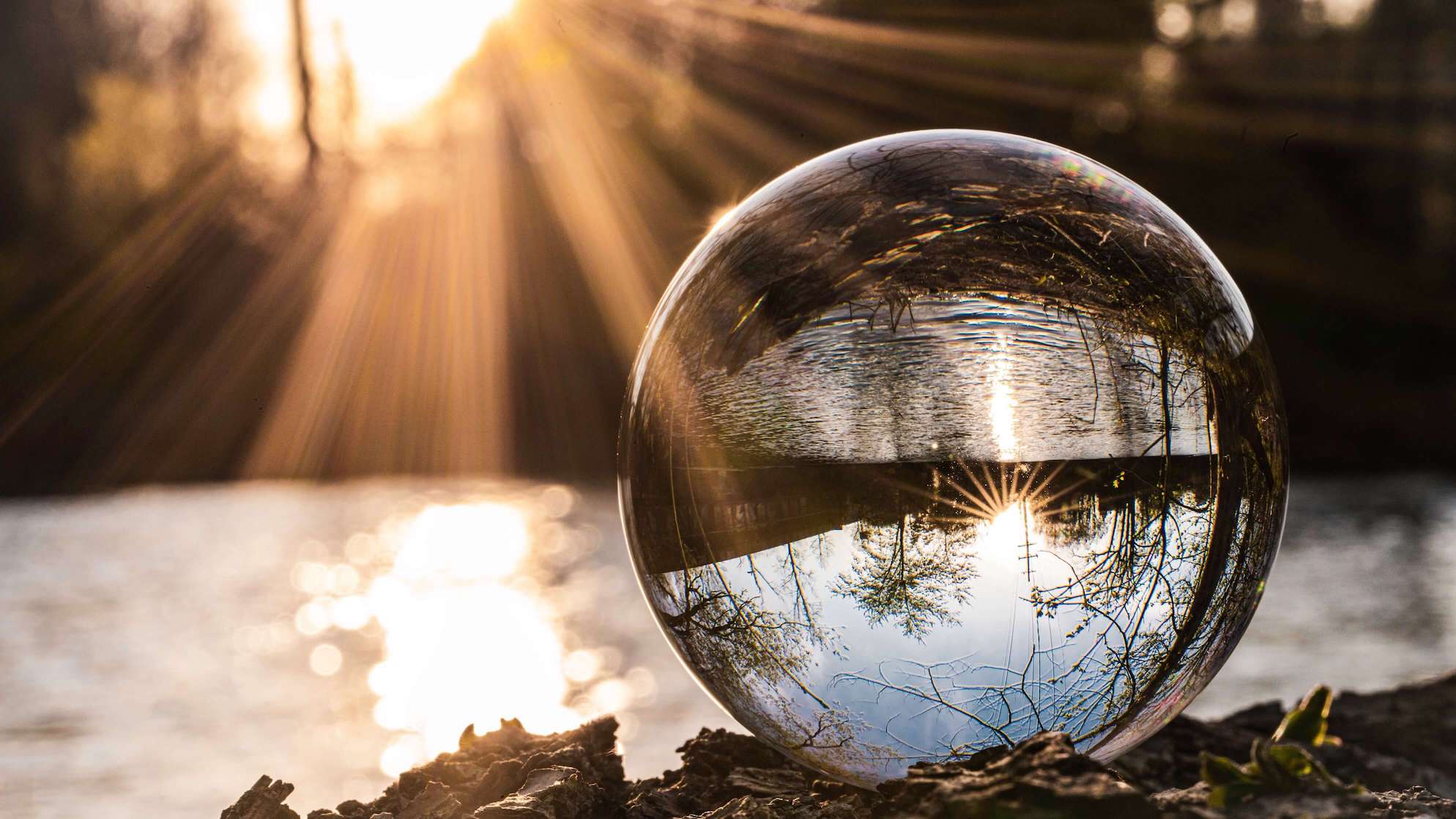 A crystal ball on a pile of leaves. It shows an upside-down reflection of a river and sunshine from the backdrop.