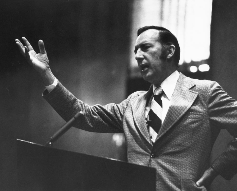 Derek Prince standing at the pulpit in church with his right hand raised, preaching to a crowd.