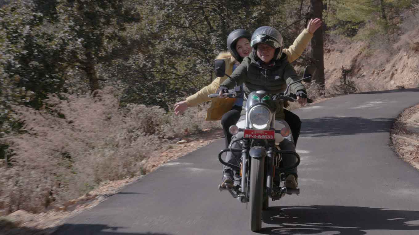 A still from "Agent of Happiness," where a man drives a motorcycle and a woman rides behind him, her hands in the air, smiling