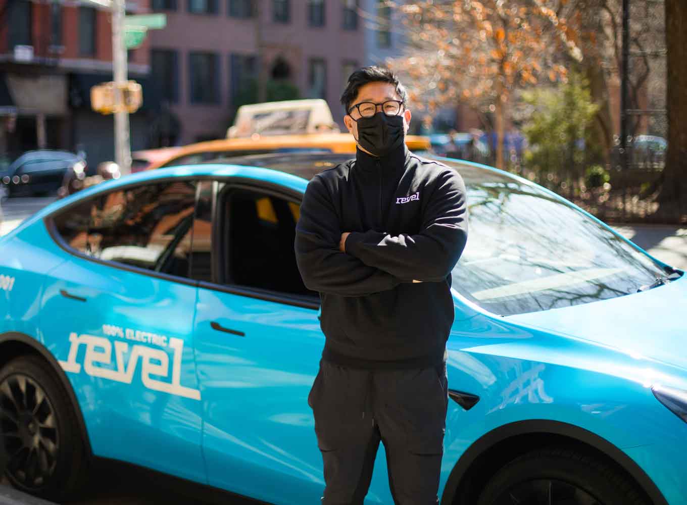 An Asian Revel driver in a black mask and glasses stands outside of a blue vehicle, his arms crossed in front of his chest