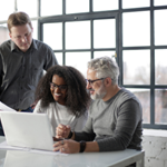 Three people stand around and talk while one is on a laptop computer.