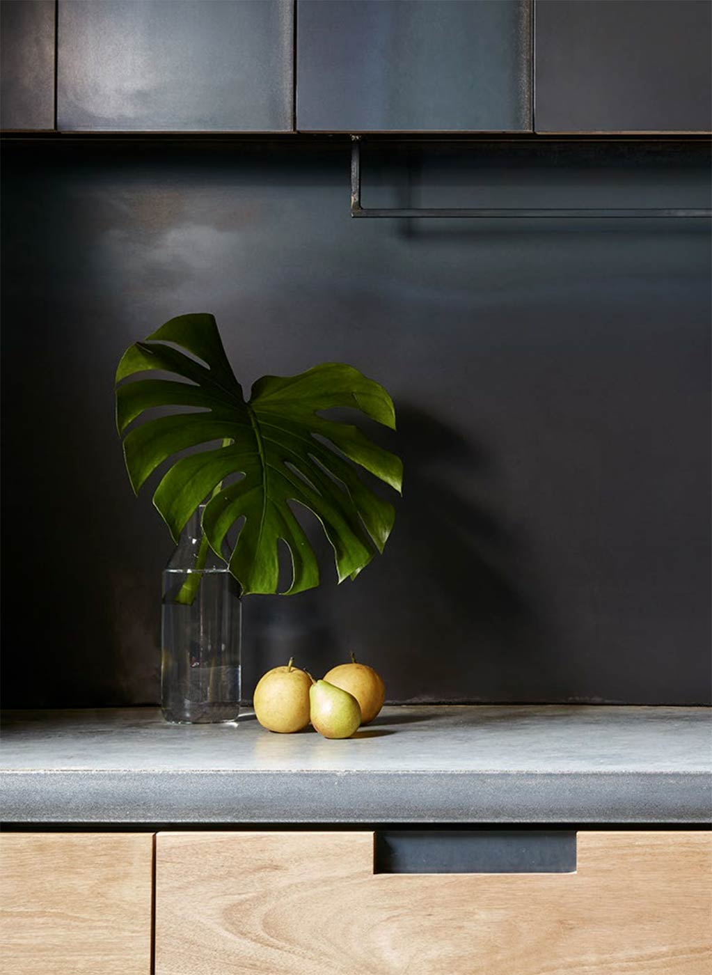 A kitchen benchtop made from concrete with wooden cabinets below. On the benchtop: some fruit and a vase with an indoor plant.