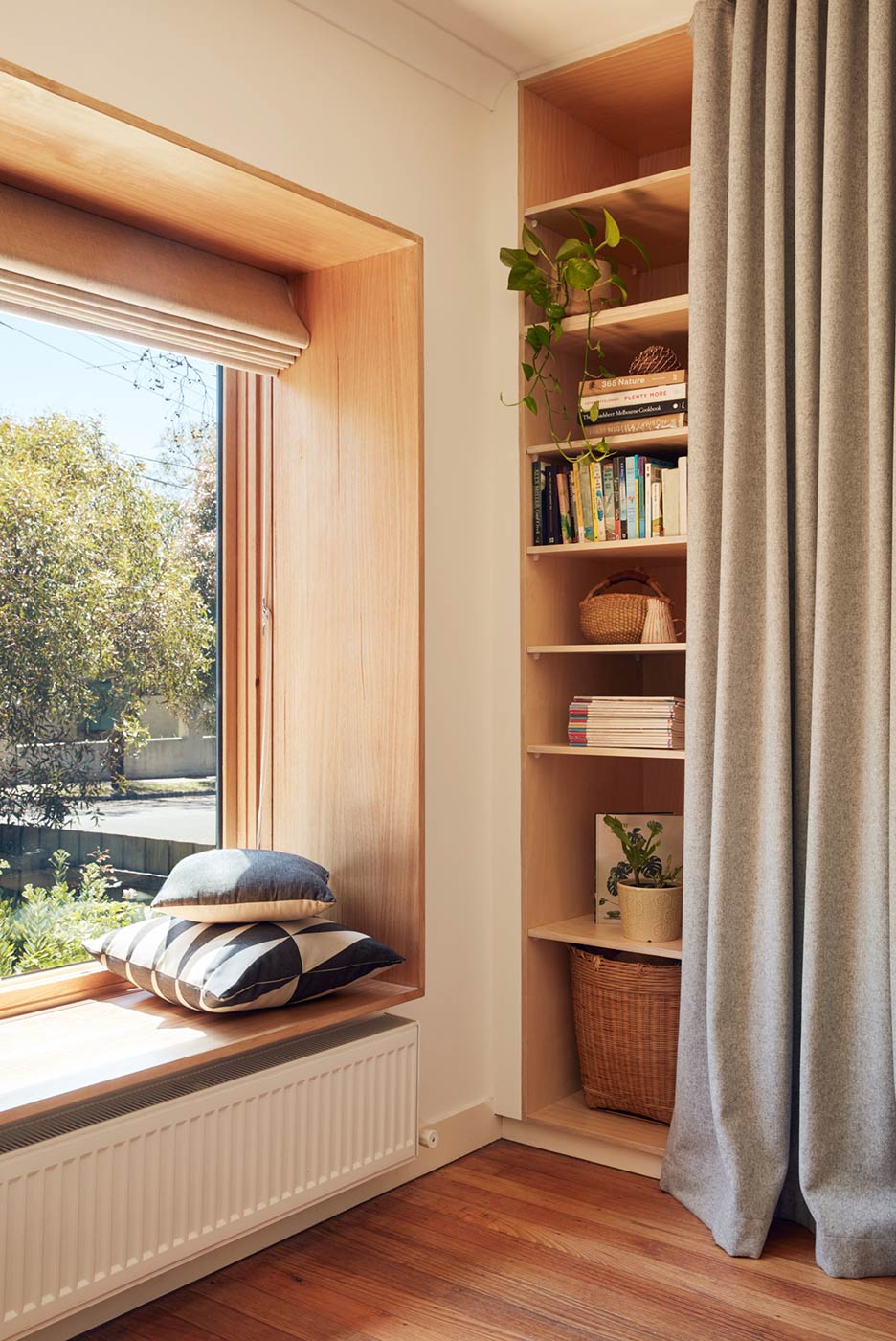 A windows with a view at the greenery outside. Next to the window: timber joinery half-covered by fabric curtain. A hydronic heating panel is installed below the window.