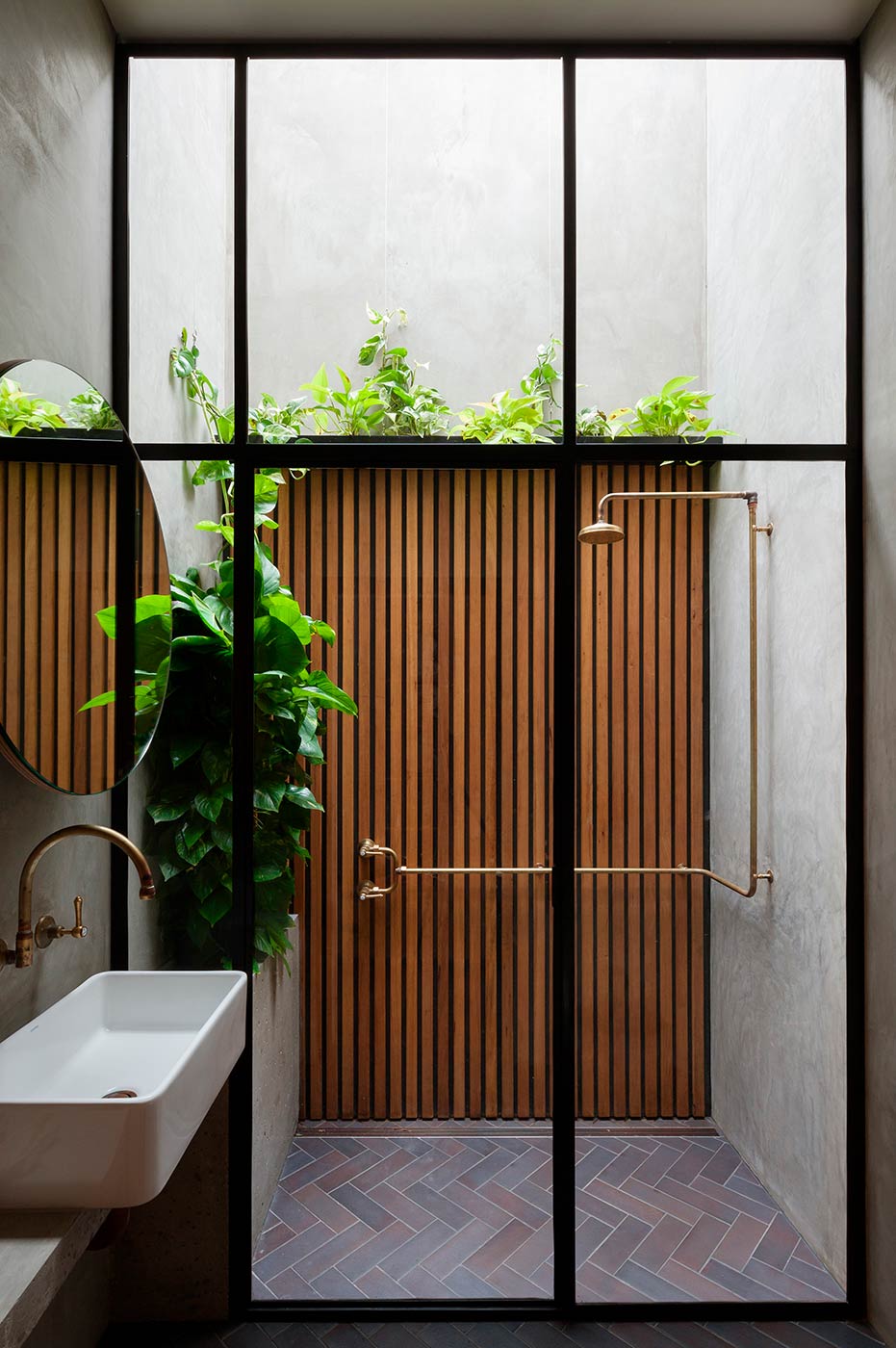 An outdoor shower in a small courtyard. Timber slats on the back wall with planting on top. Tapware made from brass. To the left, a bathroom vanity with a mirror.