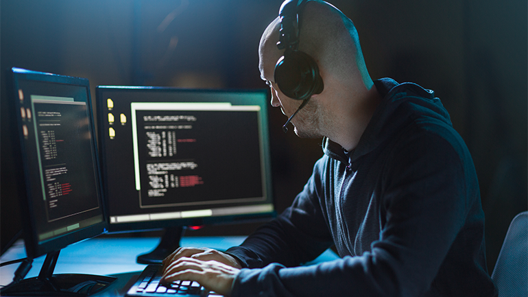 A man sits at a desk with two monitors and looks at code on the screens.