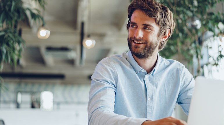 Aumente o nível de sua pilha de tecnologia de gerenciamento de conteúdo adicionando um DMS baseado em nuvem, um homem sentado em uma mesa enquanto olha para o lado direito sorrindo.