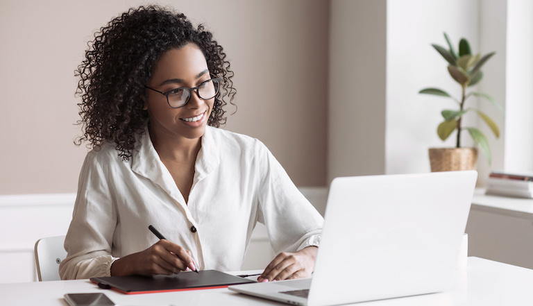 Uma mulher está sentada em uma mesa sorrindo enquanto digita em um laptop.