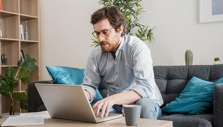 A man sits on a couch looking inventively at his laptop.