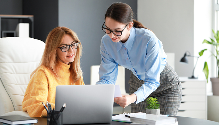 Two women sit and stand around a desk looking at a laptop screen.