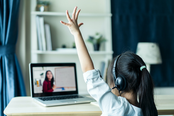 An elementary school girl attending class remotely on her laptop and raising her hand.