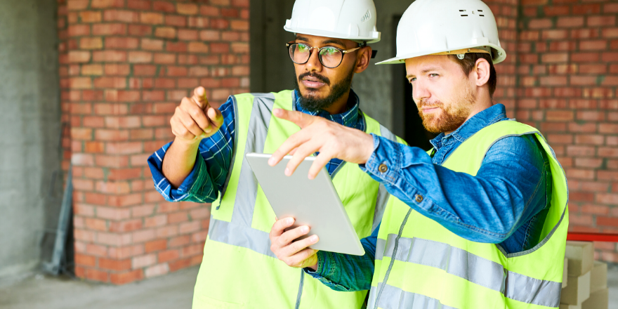 Two construction workers viewing a tablet and pointing in the distance.