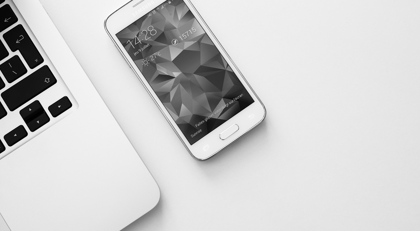 A samsung cell phone and laptop lay on a white desk top.