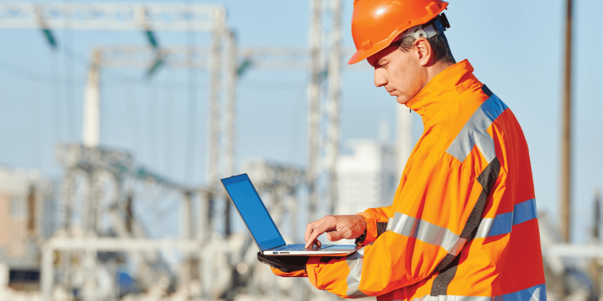 A construction worker in an orange jacket and hardhat typing on a laptop.