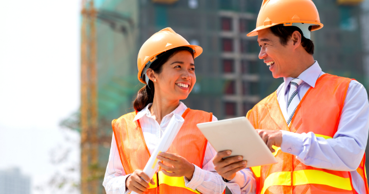 A male and female construction worker laughing and pointing at a tablet.