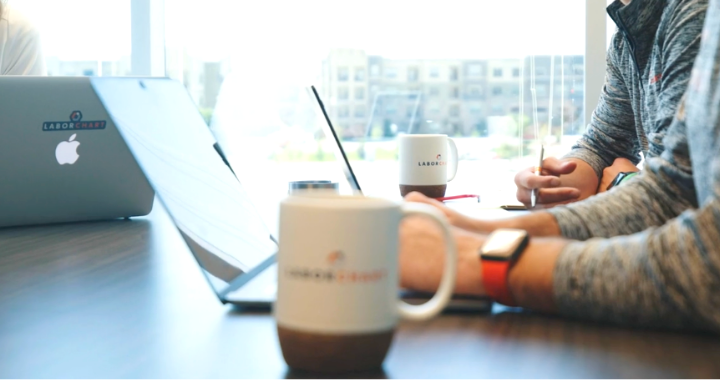 A LaborChart coffee mug with LaborChart employee's laptops and arms in the background.