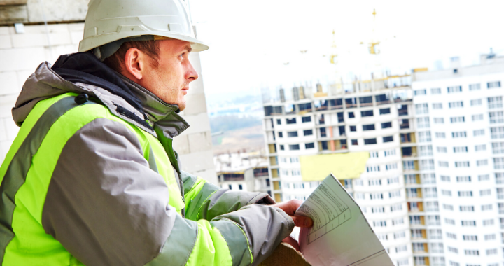 A construction worker looking off the balcony of a building holding sheets of paper.