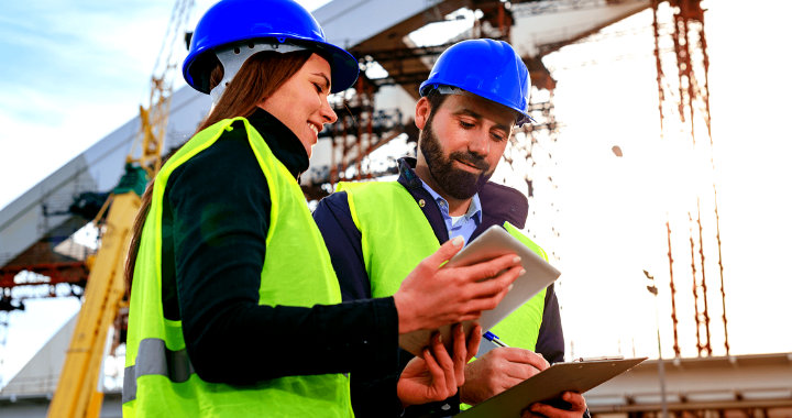 Male and female construction workers with blue hard hats managing their workforce at a construction site on a tablet.