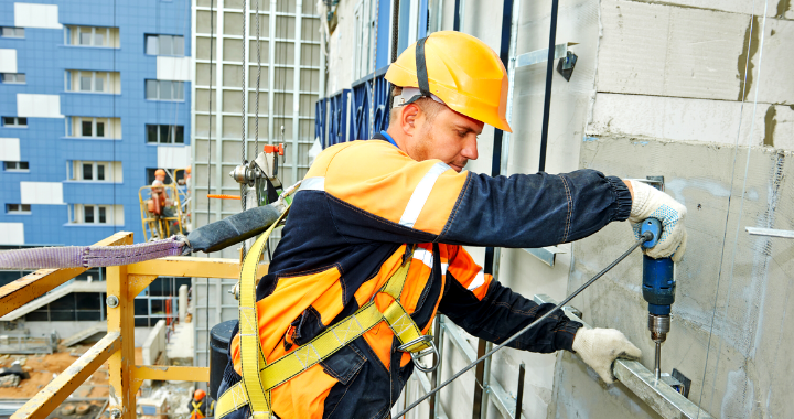 Construction worker on the side of a building drilling into a metal pole.