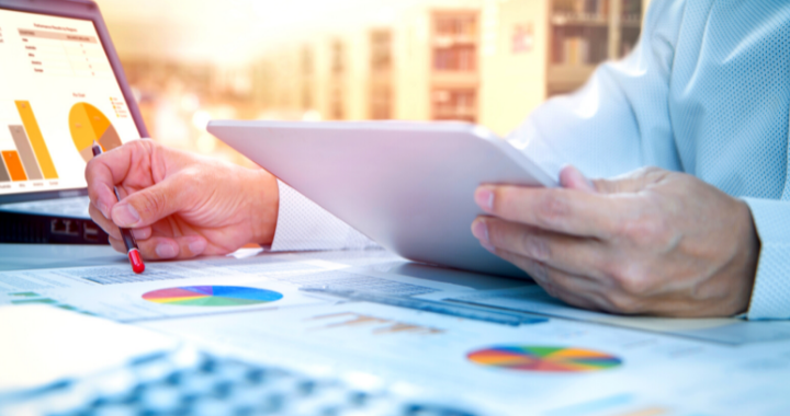 An image of a business man's desk with a tablet and pen in his hand, analyzing data all in tandem.