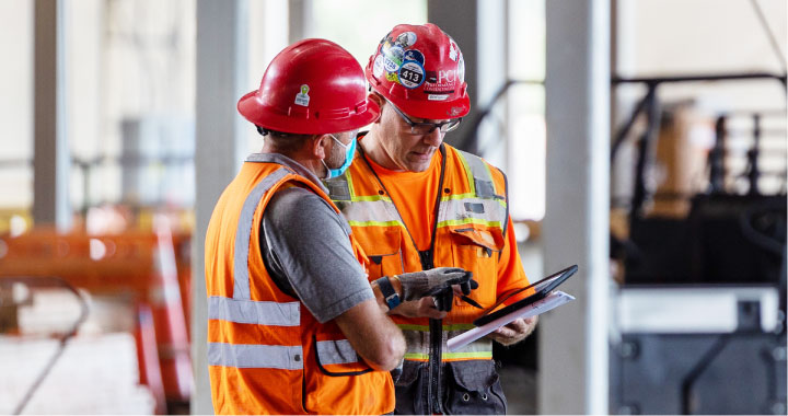 Two construction workers in red hard hats consult a tablet to manage their workforce on a jobsite.