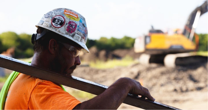 Construction worker wearing a hardhat covered in stickers carries a beam on a jobsite.