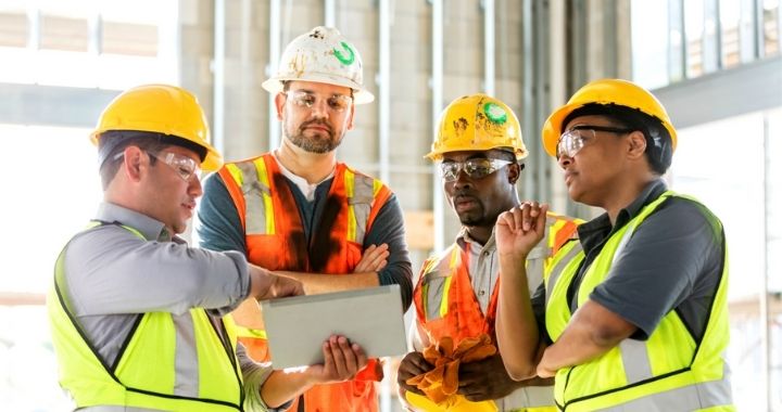 Image of four construction workers in hardhats, safety glasses and neon safety vests consult a tablet on a worksite to manage their workforce.