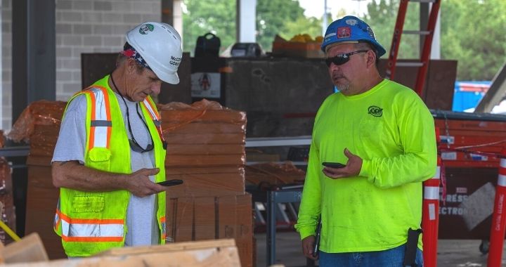 Two construction workers in a hardhats manage their workforce via their cell phones on a jobsite.