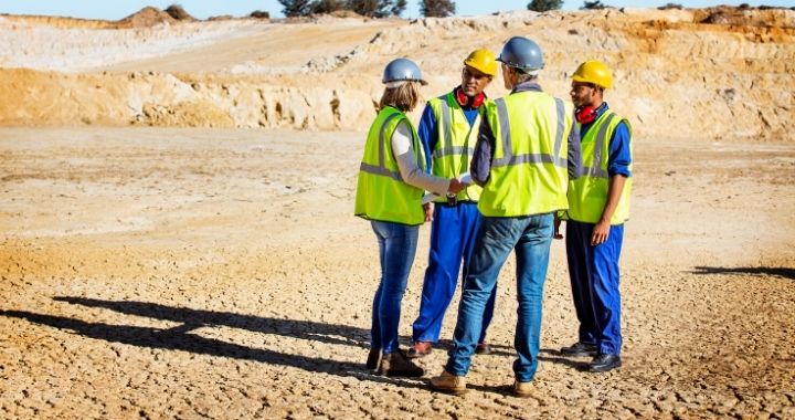 Four construction workers in hardhats, neon vests and jeans collaborate on their workforce management at a remote jobsite.