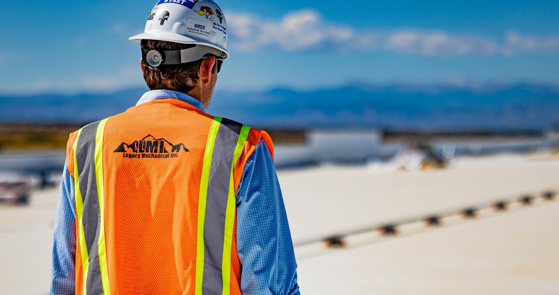 Wearing an orange vest and hardhat with stickers, a project manager with his back to the camera looks out on a project site with mountains in the distance.