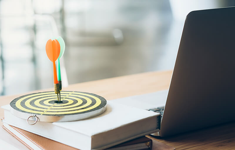 Small dartboard sitting on a desk, next to a laptop. Stuck in the center of the dartboard are a red and green dart.