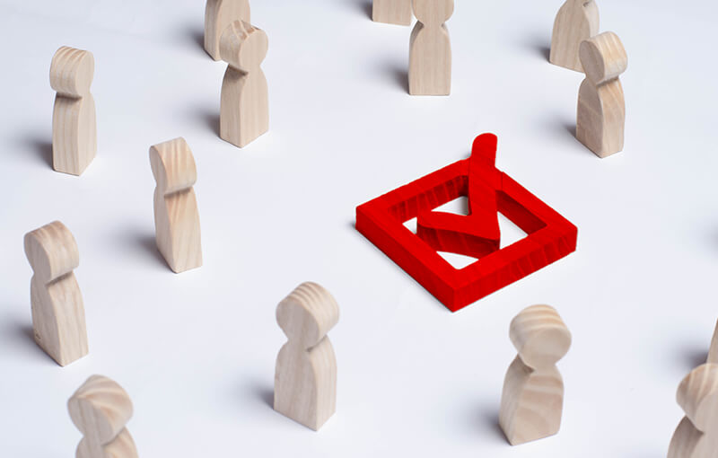 Wooden figures representing people are scattered, forming a ragged circle around a wooden red box with a wooden red checkmark in it.