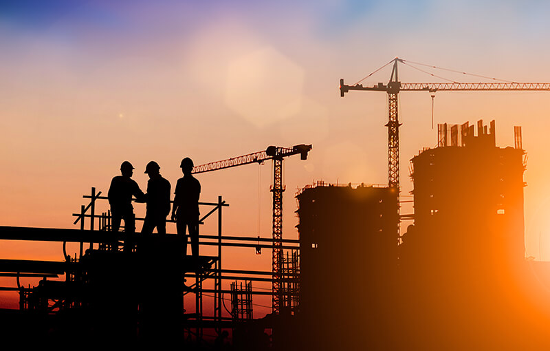 Three construction workers in front of cranes and scaffolding, silhouetted against a sunset.