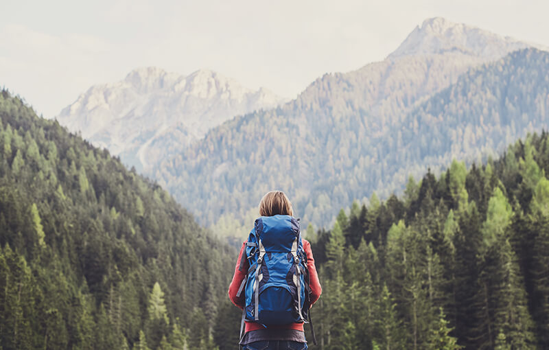 Back of a travelling woman wearing a large blue backpack, looking out over tree-covered mountains.