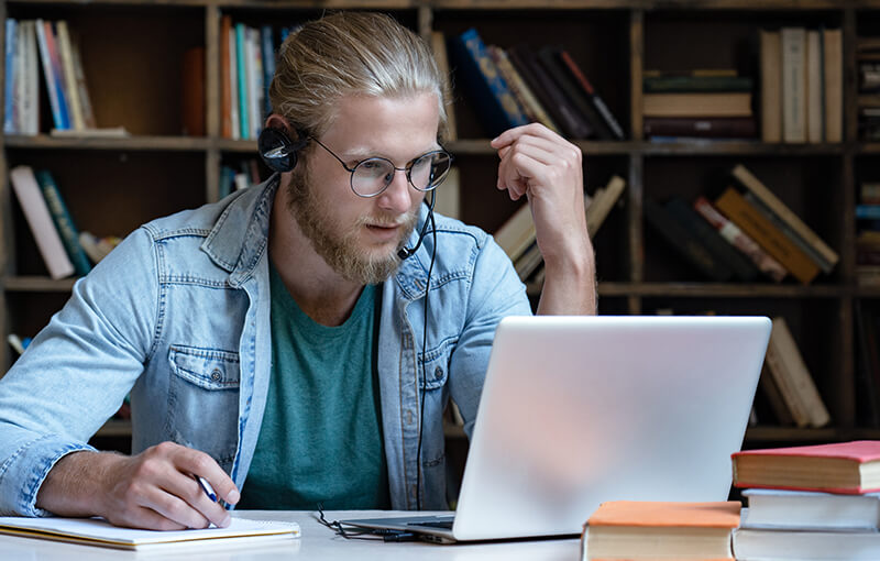 Blond, bearded man looking intently at his laptop against a backdrop of full bookshelves;.