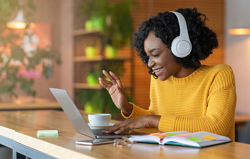 Woman with curly black hair in white headphones waving at her laptop screen.