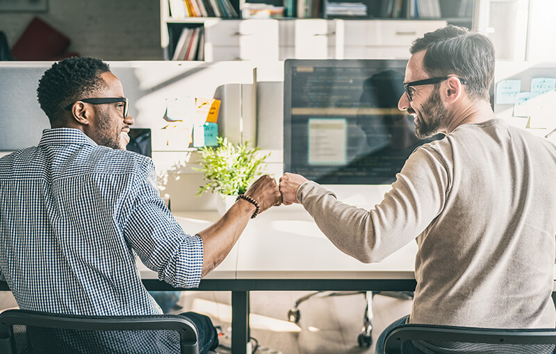 Two men sitting at a table, fist bumping.