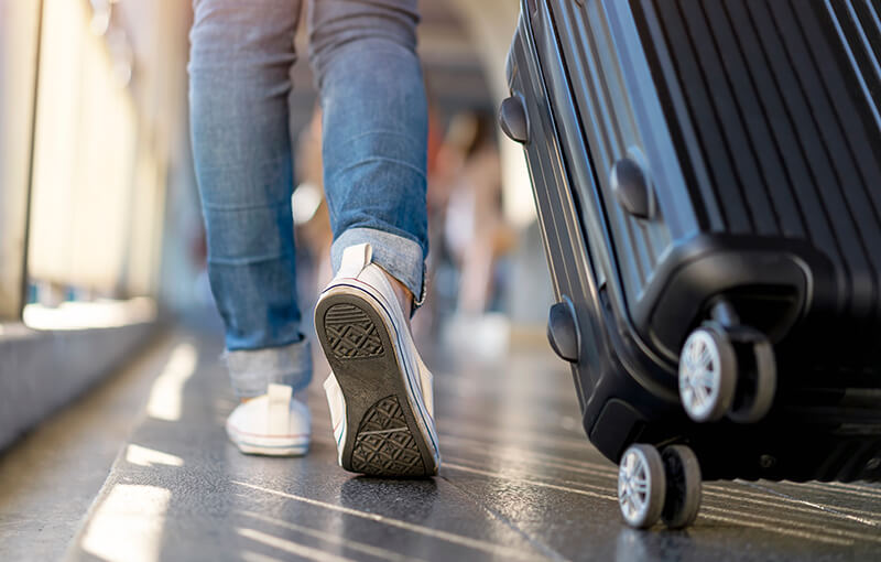 Close-up on the back of a person's legs and feet, wearing white sneakers, as they walk away, wheeling a black suitcase behind them.